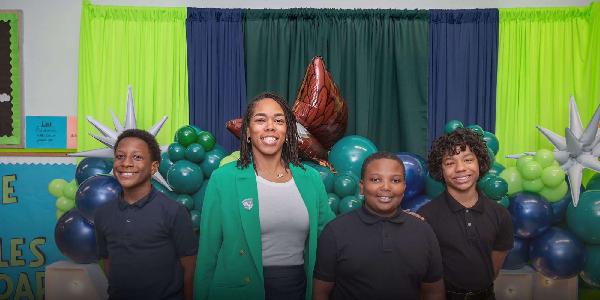 Smiling adult with three students in front of a green and blue balloon display.